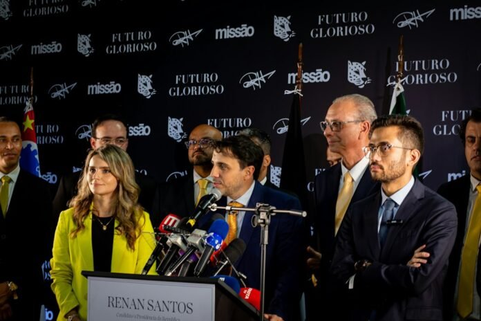 image_1790395235551 Man speaking at podium surrounded by colleagues and microphones