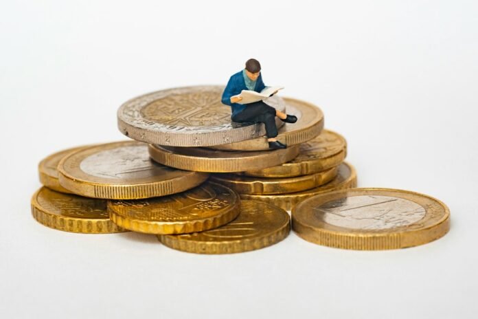 Miniature person sitting on stack of coins reading newspaper